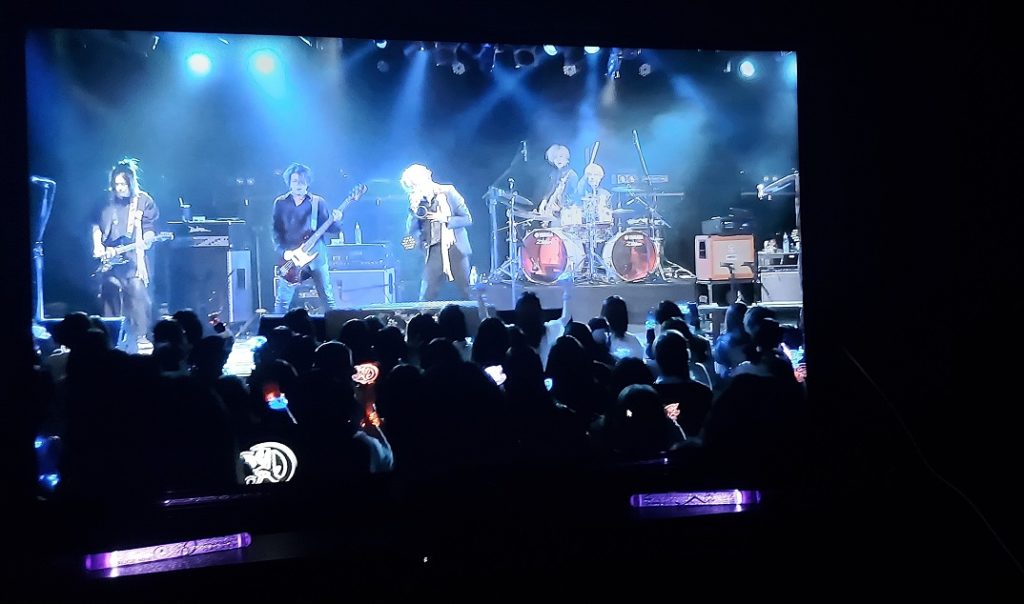 On a TV screen the five-person band ALICE NINE. performs a song a stage lit up in blue. Below the TV, two purple galaxy lights are lit as decoration.