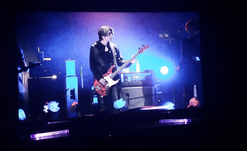 On a TV screen the focus is on Saga, playing bass; the stage lights are lit up in blue and purple. Below the TV, two purple galaxy lights are lit as decoration.