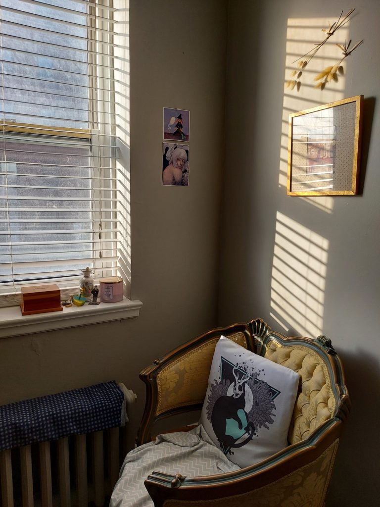 Bands of late-afternoon sunlight adorn the wall above an ornate antique chair upholstered in gold fabric. A pillow with a black cat wearing a skull with antlers on it sits on the chair. Beside it are some trinkets on a windowsill, a memorial box filled with Whiskers' ashes, a heater covered in a length of navy checkered fabric, and two prints on the wall of paintings by niyoels.