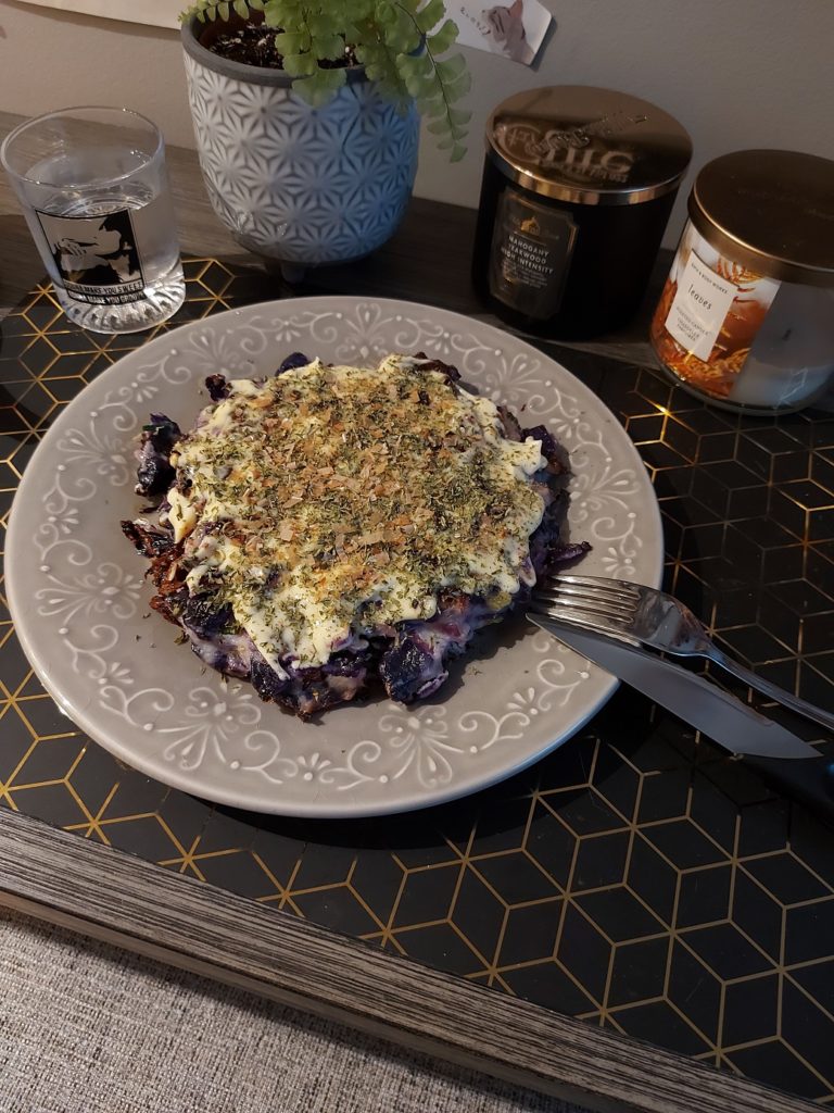 View, looking down, at a place setting with a Saga-logo glass of water and a red cabbage okonomiyaki slathered in mayonnaise, parsley flakes, and bonito flakes.