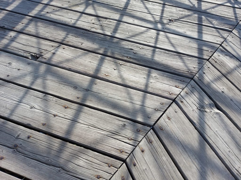 Close up of light grey wood planks studded with old nails and covered in the criss-crossing shadows of a geometric-patterned guardrail. 