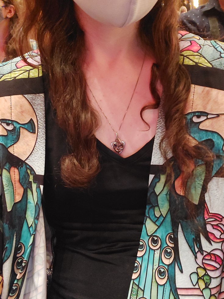 Close up of my peacock-patterned overcoat, underneath which I am wearing a black v-neck shirt and a pick-shaped silver necklace.