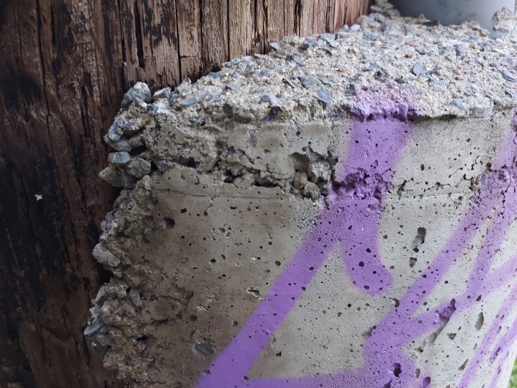 Close-up of concrete stuck to the dark wood-grain of a pole. Graffiti in purple spray-paint covers the concrete.