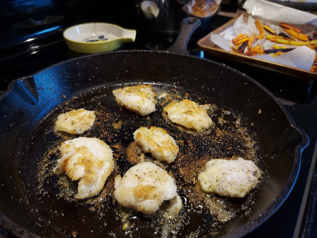 Seven cod tongues frying in a cast-iron skillet, most of them already golden-brown on the top.