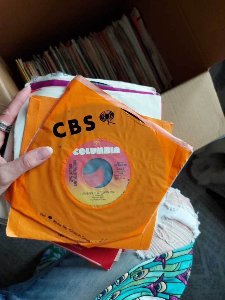 Close up of a single on vinyl; in the background, the inside of a cardboard box filled with more singles on vinyl is visible.