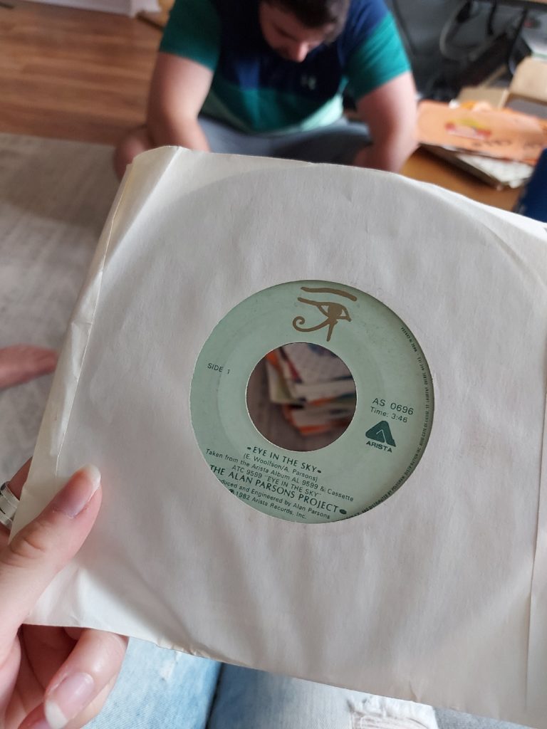 Close up of a single on vinyl; in the background, my younger brother is seated on the floor, looking through other vinyls