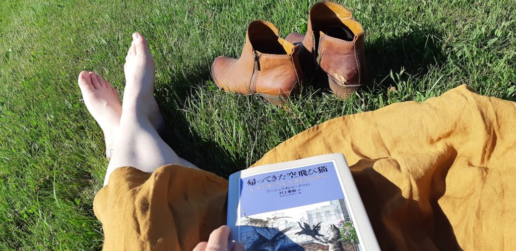 Close up of my legs folded on some grass, lit up in bright, direct sunlight. My mustard-yellow skirt is fanned out in the grass, while I hold a book in my lap: the Japanese edition of "Catwings Return" by Ursula K LeGuin. Before me, a pair of well-worn brown cowboy ankle boots sit at the ready in the grass. 