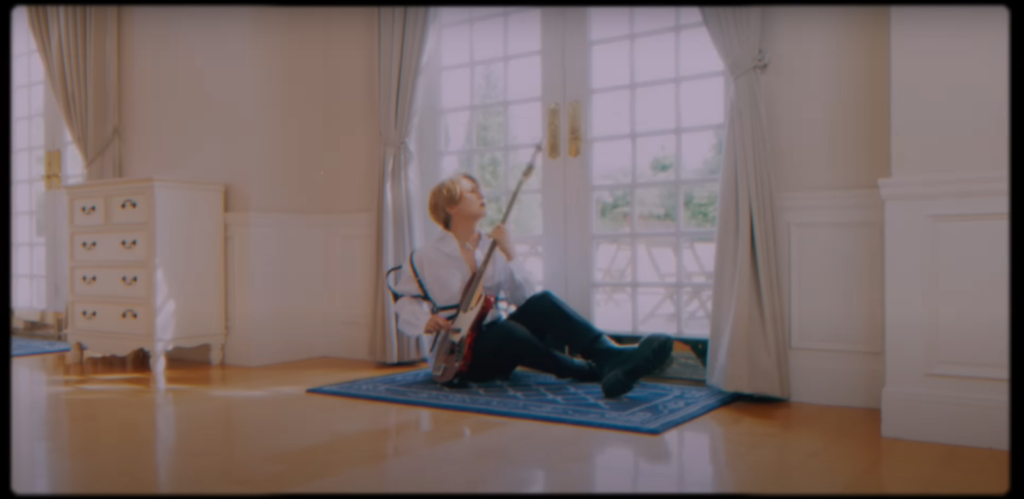 Saga playing bass in front of windowed double doors, on a carpet.