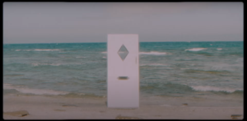 A white door with a mail slot and a diamond-shaped window stands in a beach before an ocean in daylight, waves rolling in.